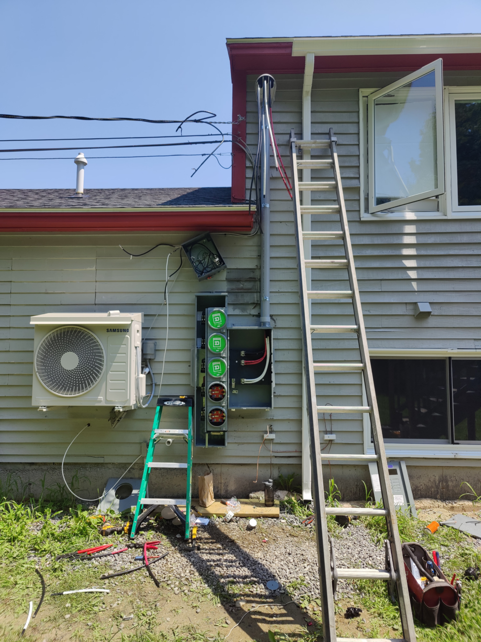 Exterior of a house with ladders leaning against the wall. An HVAC unit and electrical panel are visible, with tools and cables scattered on the ground.