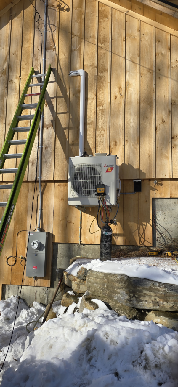 A wall-mounted outdoor unit of a heat pump system is installed on a wooden building. A green ladder rests against the wall, and snow covers the ground and nearby stones.